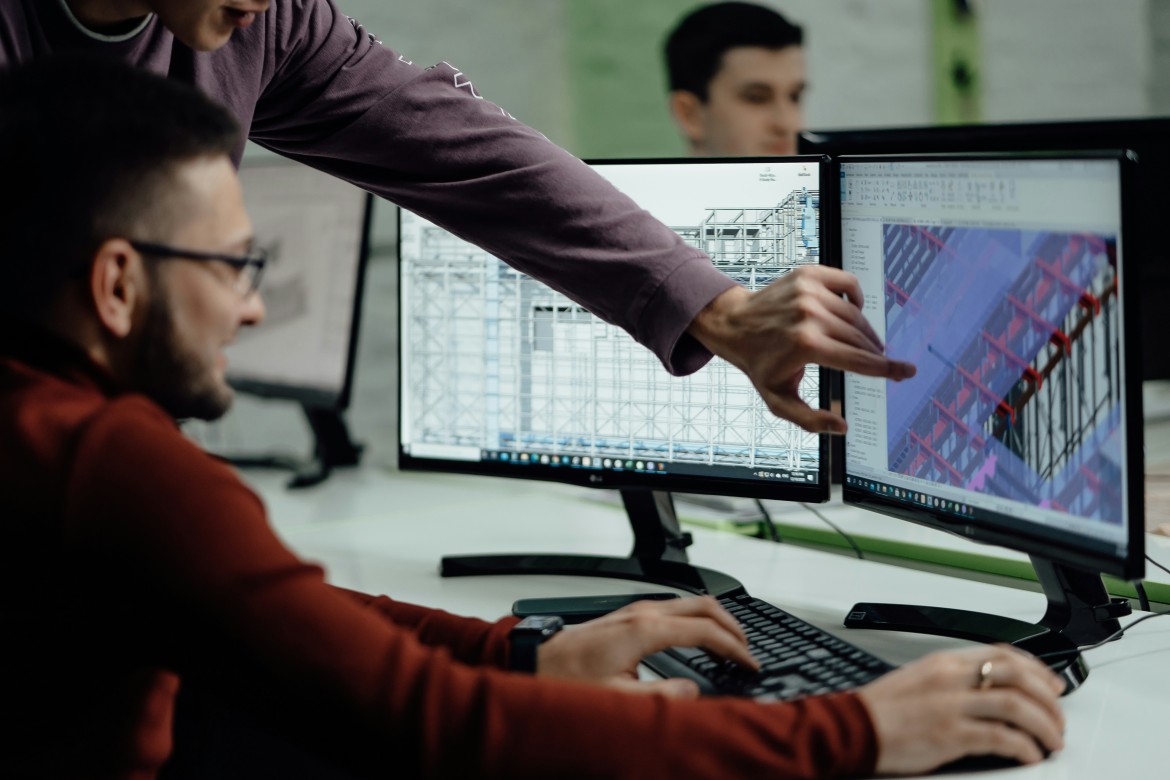 Two people collaborating at dual monitors displaying a detailed 3D wireframe and colored structural model of a building.