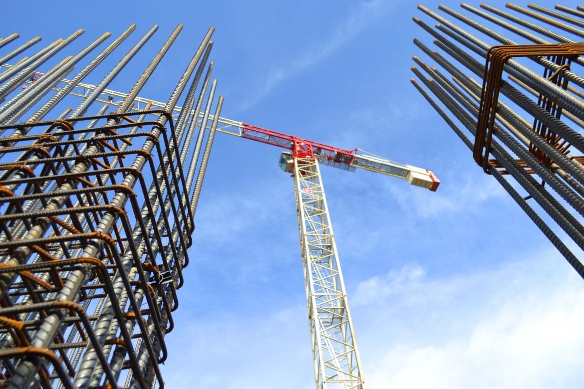 Tower crane above reinforced steel bars at a construction site, viewed from below against a blue sky.